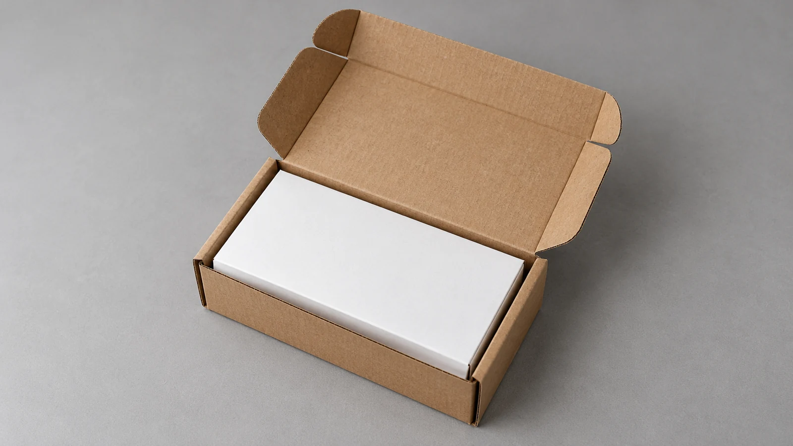 An open brown corrugated mailer closely fitted around a plain white product carton on a clean grey surface.